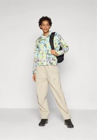 Γυναίκα που φοράει παστέλ παραλλαγής μπουφάν, μπεζ cargo παντελόνια, μαύρες πεζοπορικές μπότες και κρατάει μια μαύρη σακίδιο στους ώμους.