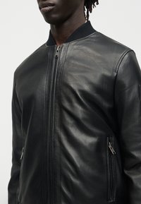 Κοντινό πλάνο ατόμου που φοράει μαύρο δερμάτινο μπουφάν bomber με φερμουάρ μπροστά και φερμουάρ στις πλαϊνές τσέπες, μπροστά σε απλό φόντο.