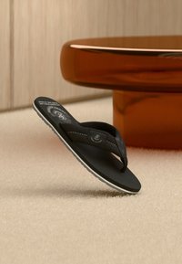 Μαύρο σαγιονάρι με λευκή σόλα και λουράκι με λογότυπο, τοποθετημένο όρθιο σε μπεζ χαλί δίπλα σε στρογγυλό καφέ τραπέζι.