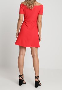 Vestido rojo de manga caída con cintura acanalada y bajo acampanado, combinado con sandalias negras de tacón alto que tienen tiras de lunares y tacones gruesos.