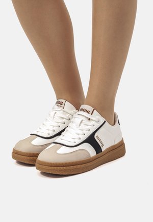 Zapatillas blancas y beige con acentos negros, suela de goma texturizada, cierre de cordones y un logo en la lengüeta. Materiales de ante y sintéticos utilizados.