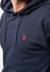 Dunkelblaues Hoodie aus weichem Stoff, mit Kapuze und Kordelzug, strukturierten Details und einem roten Logo, das auf der Brust gestickt ist.