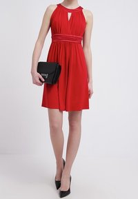 Robe rouge sans manches avec un décolleté en trou de serrure et une taille cintrée. Complétée par une pochette noire et des talons hauts noirs. Tissu lisse.