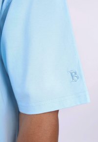 Lys blå t-skjorte med korte ermer laget av bomull, med et lite brodert bokstav "B" på ermet og en glatt tekstur.