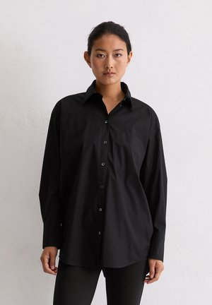 Woman with dark hair tied back wears loose black button-up shirt and black pants against plain light wall background.