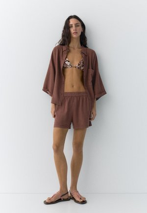 Femme debout contre un mur blanc, portant un haut de bikini à motifs, une chemise marron ample ouverte, un short assorti et des sandales roses à brides.