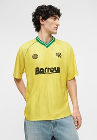 Joven con cabello rizado que lleva una camiseta amarilla Barrow con logotipos verdes y negros, combinada con jeans azul claro, de pie contra un fondo liso.