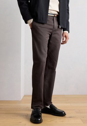 Mann trägt dunkelbraune, gerade geschnittene Hose, schwarze Slipper mit weißen Socken, eine schwarze Jacke und einen beigefarbenen Pullover, steht auf einem Holzboden.