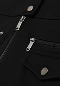 Abrigo negro con cremalleras metálicas, bolsillos de presión y un tejido texturizado con un acabado suave. Los acentos incluyen herrajes plateados.