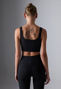 Kvinne med håret i en bun som vender bort, iført svarte sportsklær, og viser en detaljert tatovering som dekker hennes øvre rygg og skuldre.