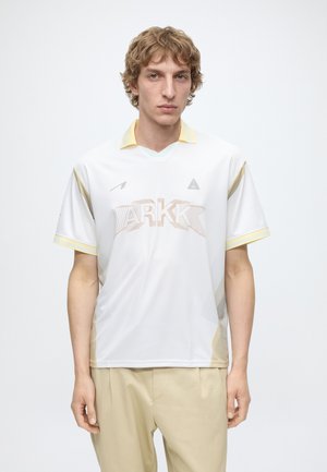 Junger Mann mit hellbraunen lockigen Haaren, trägt ein weißes ARKK-Sportshirt und beige Hose, steht vor einem schlichten weißen Hintergrund.