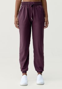 Bordeauxfärgade joggerbyxor med dragsko i midjan, sidofickor och elastiska cuffs. Har en mjuk, lätt tyg. Bärs med vita skor.