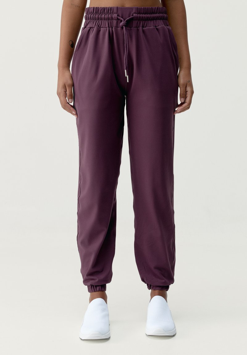 Bordeauxfärgade joggerbyxor med dragsko i midjan, sidofickor och elastiska cuffs. Har en mjuk, lätt tyg. Bärs med vita skor.