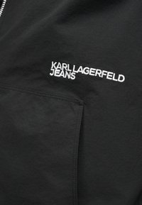 Μαύρο ύφασμα με λευκό κεντημένο λογότυπο "KARL LAGERFELD JEANS" πάνω από λεπτομέρεια ραμμένης τσέπης.
