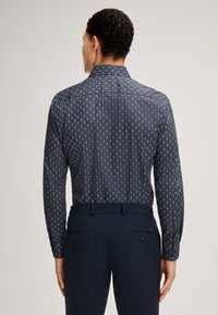 Camicia blu navy con motivo a piccoli disegni bianchi, maniche lunghe e colletto strutturato, abbinata a pantaloni scuri con tasche posteriori.