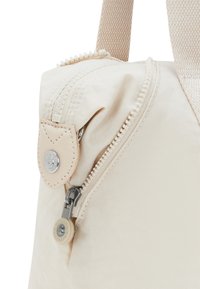 Beige Stoffhandtasche mit Reißverschluss, ausgestattet mit einem strukturierten Tragegriff, metallischen Beschlägen und einem runden Logo-Akzent am Reißverschluss.