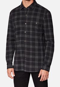 Man in een zwart en donkergrijs geblokt overhemd met lange mouwen en een boord, gecombineerd met zwarte broeken.