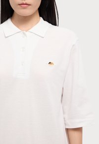 Polo majica od svijetlo ružičastog pamuka, s tri dugmeta na izrezu, kratkim rukavima i malim izvezenim logotipom na lijevoj strani prsnog koša.