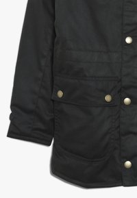 Czarna materia�łowa kurtka z rękawem i kieszenią z metalowymi napami z przodu, prezentująca teksturowane przeszycia i gładki materiał.