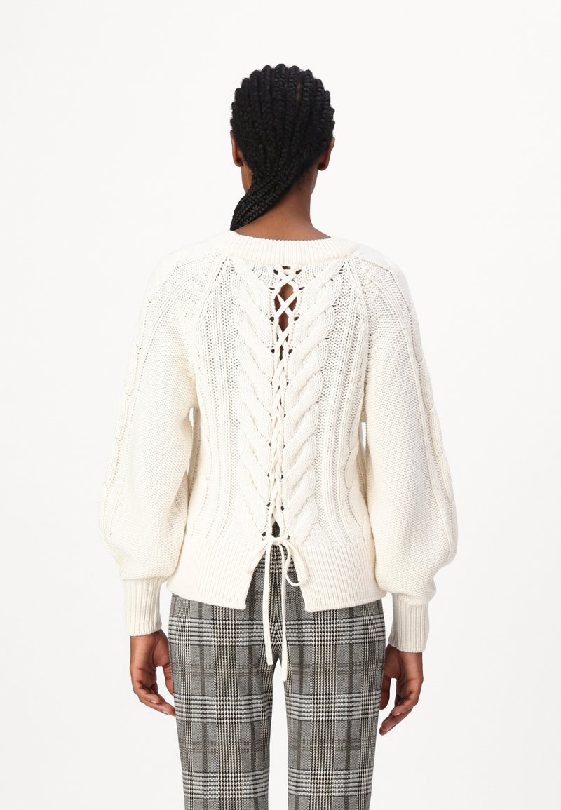 Femme aux cheveux tressés portant un pull en maille torsadée blanc avec un détail à lacets dans le dos et un pantalon à carreaux gris, de dos.