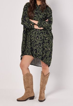 Vrouw die een oversized groene en zwarte jurk met luipaardprint draagt, met opgerolde mouwen en lichtbruine suède kniehoge laarzen, staand met gekruiste armen.