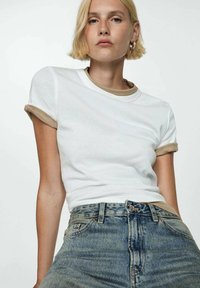 T-shirt bianco a maniche corte con risvolti color sabbia, indossato sopra una maglietta girocollo beige, abbinato a jeans in denim a vita alta.