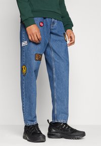 Denimjeans med avslappnad passform, blå färg, med olika broderade lappar som symboler och text. Bärs med svarta vandringsskor.