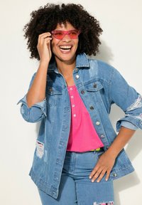 Giacca di jeans con dettagli usurati, colore blu chiaro, chiusura a bottoni, abbinata a una camicia rosa e jeans blu. Occhiali da sole rosa a farfalla.