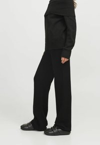 Zwarte oversized gebreide trui met off-shoulder ontwerp, gecombineerd met zwarte wijde broek en zwarte instapschoenen. Zachte textuur, minimale details.
