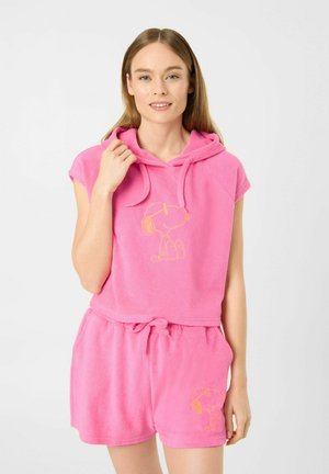 Jeune femme portant un sweat à capuche sans manches rose et un short avec un motif Snoopy, souriant devant un fond blanc uni.