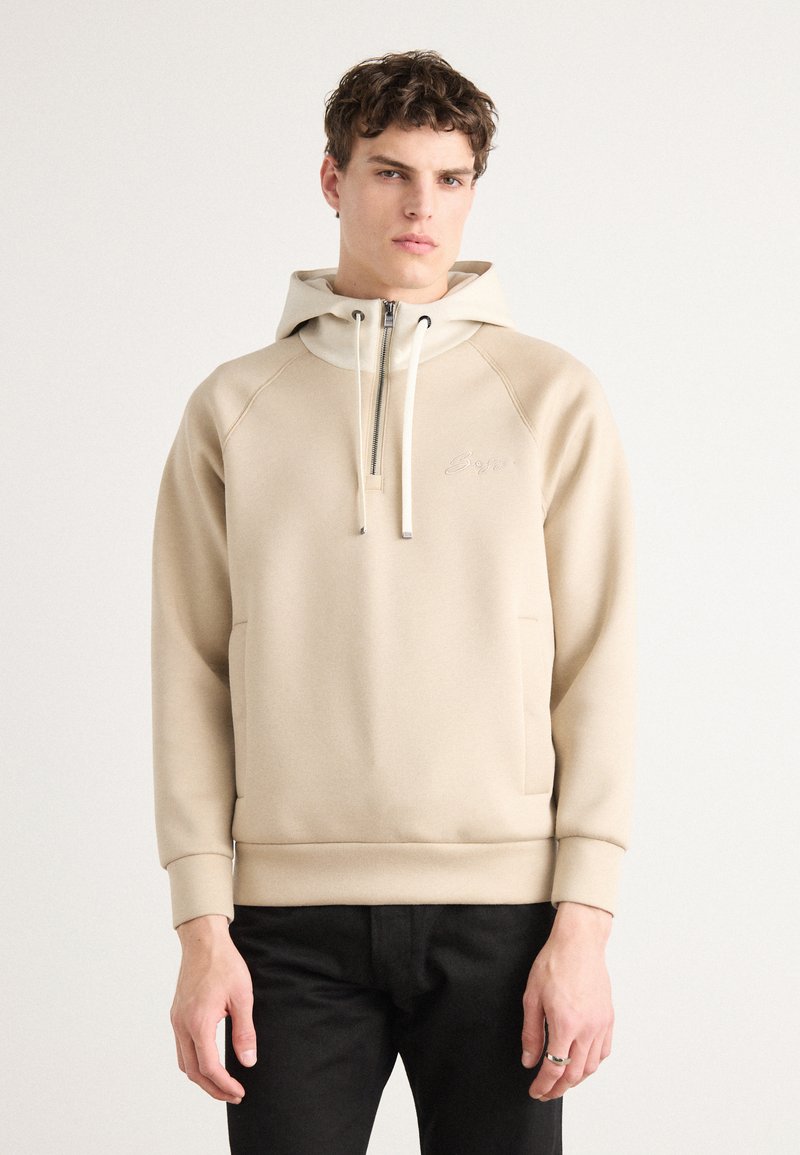 Sweatshirt à capuche beige avec un col zippé, deux poches avant, des poignets côtelés et un logo brodé sur le devant. Tissu doux et texturé.