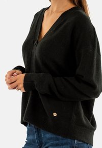 Cardigan de lana negro con escote en V, mangas largas y una forma ligeramente oversized. Presenta un pequeño acento de marca cerca del dobladillo.