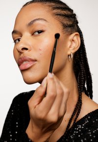Pennello per trucco con setole nere tenuto in mano, mentre si applica il prodotto sulla guancia. Il modello ha i capelli intrecciati e indossa un top di paillettes nero. La pelle ha un naturale splendore.