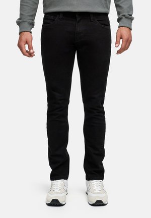 Mann trägt schlanke schwarze Jeans und weiße Sneakers, steht mit entspannten Armen seitlich an einem schlichten Hintergrund.