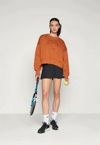 Kobieta w pomarańczowej bluzie i czarnej spódnicy tenisowej trzyma rakietę tenisową i piłkę, ubrana w czarne buty sportowe i białe skarpetki, na tle jednolitego tła.