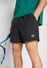 Pantalones cortos deportivos negros con una textura suave, una cintura elástica y un logo en la parte inferior izquierda. Combinados con una camiseta verde.