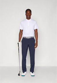 Golfklädsel med en figursydd vit pikétröja, marinblå byxor, grått bälte, vita skor med blå detaljer och en golfklubba i handen.