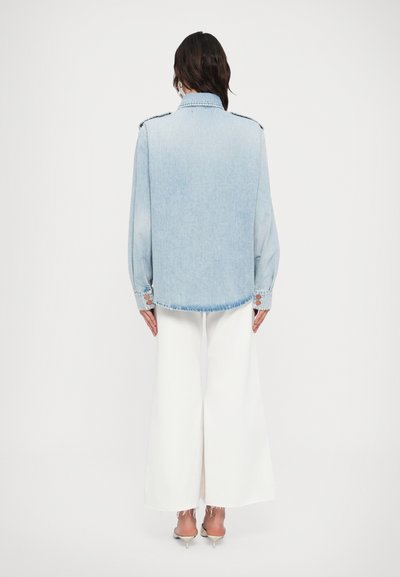 Τζιν μπουφάν σε ανοιχτό μπλε χρώμα με χαλαρή εφαρμογή, μανσέτες με κουμπιά και κλασικό γιακά. Συνδυασμένο με φαρδιά λευκά παντελόνια. Πλάγια όψη από πίσω.