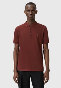 Polo shirt marrone con colletto, dotato di tre bottoni e un piccolo logo. Indossato con pantaloni neri, su uno sfondo neutro.