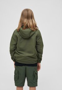 Veste à capuche verte avec des poignets et une taille élastiques. Associée à un short cargo vert, le tout en tissu lisse et résistant. Vue arrière affichée.