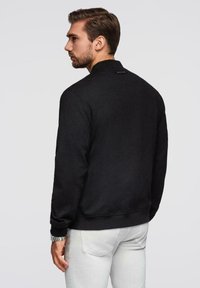 Czarny sweatshirt z wysokim kołnierzem, teksturowana tkanina i ściągacze przy mankietach. Zawiera małą metkę z logo na plecach. Noszony z jasnymi spodniami.