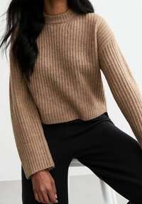 Pull en maille côtelée brun avec un col haut et des manches larges, présentant un design court. Porté avec un pantalon noir. Texture lisse.