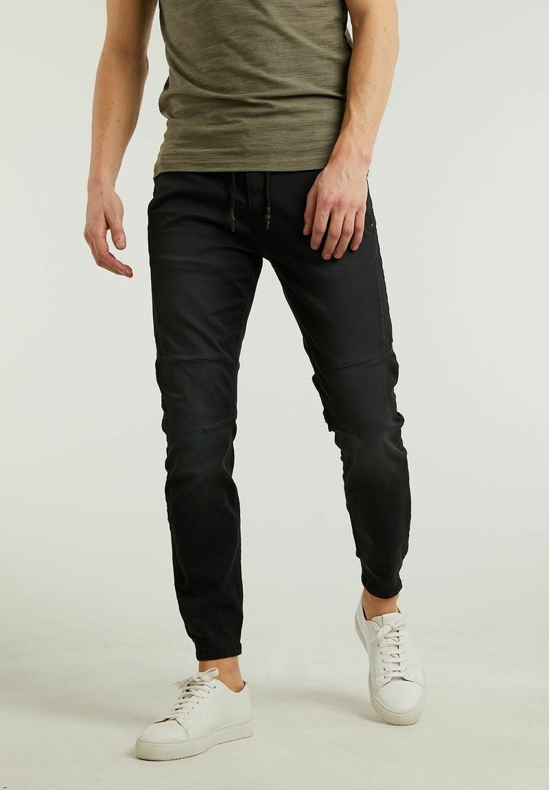 Homme portant un pantalon slim noir, un haut à manches courtes vert olive et des baskets blanches, debout devant un fond uni.