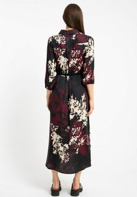 Kobieta stojąca plecami do kamery, ubrana w długą czarną suknię z maroonowymi i beżowymi wzorami kwiatowymi oraz czarne platformy.