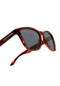 Ochelari de soare Hawkers cu o ramă lucioasă de tortoise, lentile întunecate și un design cat-eye curbat. Prezintă un logo argintiu pe tâmplă.