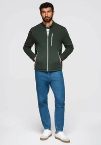 Grön zip-up jacka med hög krage, texturerat tyg, sidofickor med dragkedja, kombinerad med blå jeans och vita sneakers. Modellen står med händerna i fickorna.