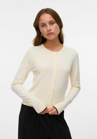 Crèmekleurige knoopcardigan met lange mouwen, ribbelige randen en een aansluitend ontwerp, gecombineerd met een geplooide zwarte rok.