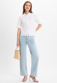 Witte kortlijfknopshirt, lichtblauwe high-waisted jeans, zilveren hoge hakken, met een geweven straw handtas.