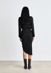Schwarzes Langarmshirt, schmale schwarze Midirock mit asymmetrischem Saum, kombiniert mit schwarzen Absatzstiefeln. Glatte Textur, minimalistische Hardwaredetails.