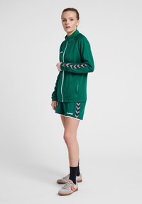 Groene sportjas met een gestructureerde afwerking, witte rits en grijze chevron-accenten op de mouwen, gecombineerd met bijpassende shorts. Grijze sportieve schoenen.